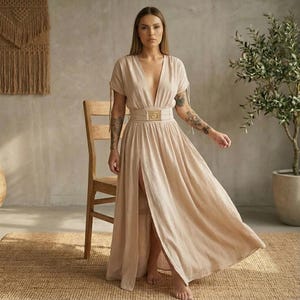 May include: A beige maxi dress with a plunging neckline and a high slit. The dress features short sleeves with tie details and a wide belt with a gold-colored buckle. The dress is made of a flowing fabric, suitable for warm weather.