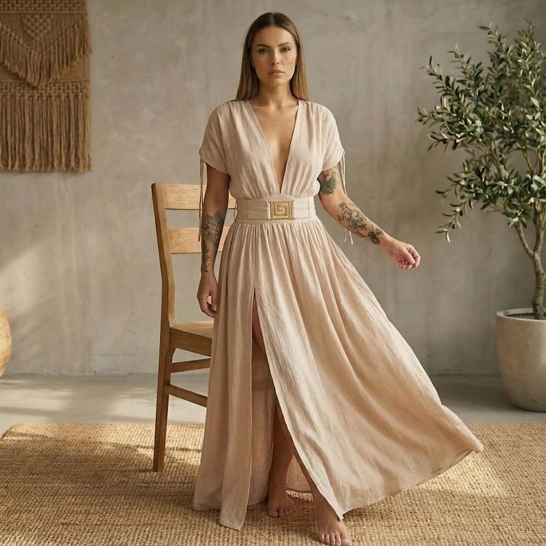 May include: A beige maxi dress with a plunging neckline and a high slit. The dress features short sleeves with tie details and a wide belt with a gold-colored buckle. The dress is made of a flowing fabric, suitable for warm weather.