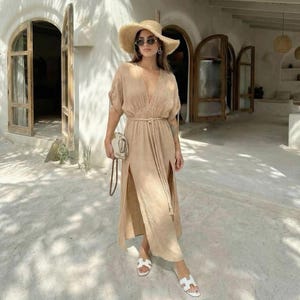 Puede incluir: Vestido largo beige con cuello en V, mangas cortas y aberturas laterales. El vestido est&aacute; ce&ntilde;ido a la cintura con un cintur&oacute;n de cuerda a juego. La persona lleva un sombrero de paja, gafas de sol, sandalias blancas y un peque&ntilde;o bolso blanco roto.