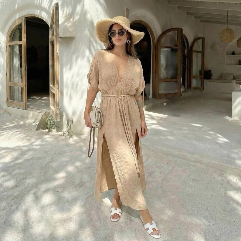 Puede incluir: Vestido largo beige con cuello en V, mangas cortas y aberturas laterales. El vestido est&aacute; ce&ntilde;ido a la cintura con un cintur&oacute;n de cuerda a juego. La persona lleva un sombrero de paja, gafas de sol, sandalias blancas y un peque&ntilde;o bolso blanco roto.
