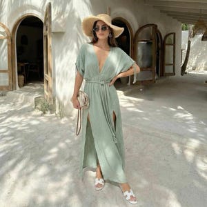 Puede incluir: Vestido largo verde claro con escote pronunciado y aberturas laterales altas. El vestido se ci&ntilde;e a la cintura con un cintur&oacute;n de cuerda a juego. La persona lleva un sombrero de paja, gafas de sol y sandalias blancas. Se sostiene un peque&ntilde;o bolso beige en la mano.