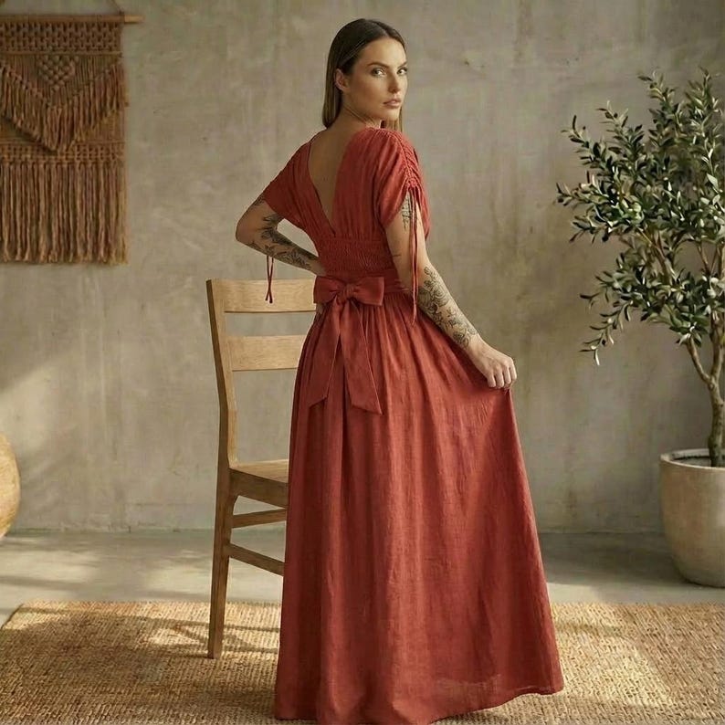 May include: A rust-colored maxi dress with a deep V-neck and short sleeves. The dress features a bow at the waist and a flowing skirt. The dress is made of a lightweight fabric, perfect for warm weather.