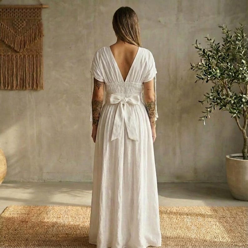 May include: A white maxi dress with a V-neckline and short sleeves. The dress features a smocked waist and a large bow in the back. The dress is made of a lightweight fabric.