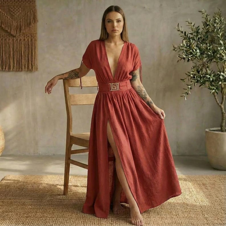 May include: A rust-colored maxi dress with a deep V-neckline and short sleeves. The dress features a high slit and is cinched at the waist with a decorative belt. The dress is made of a flowing fabric.