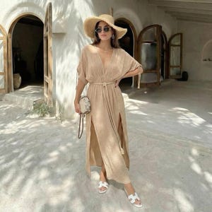 Puede incluir: Vestido largo beige con cuello en V, aberturas laterales y cintur&oacute;n de cuerda. El vestido se combina con un sombrero de paja, gafas de sol y sandalias blancas. Un peque&ntilde;o bolso estampado se sostiene a un lado.