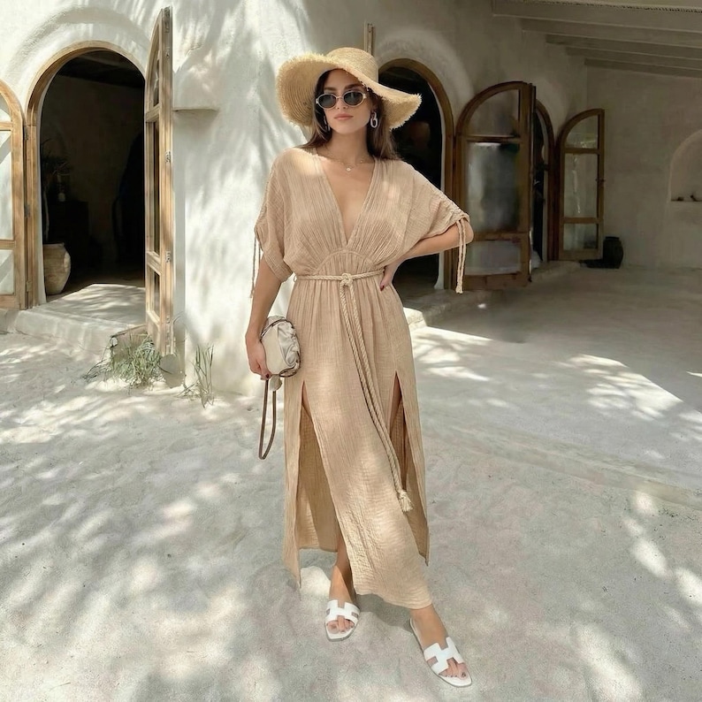 Puede incluir: Vestido largo beige con cuello en V, aberturas laterales y cintur&oacute;n de cuerda. El vestido se combina con un sombrero de paja, gafas de sol y sandalias blancas. Un peque&ntilde;o bolso estampado se sostiene a un lado.