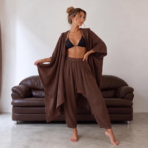 May include: A dark brown two-piece set consisting of a hooded, open-front poncho with wide sleeves and matching high-waisted, loose-fitting trousers. The fabric has a textured, crinkled appearance. A black triangle bikini top is worn underneath.