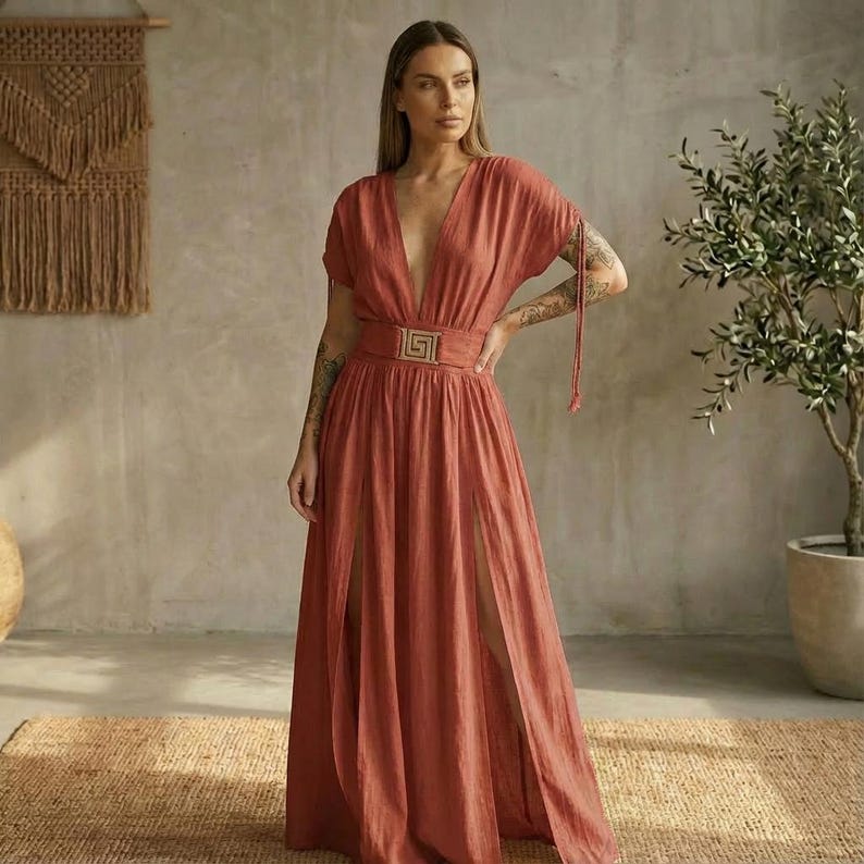 May include: A rust-colored maxi dress with a plunging neckline and short sleeves. The dress features a decorative belt with a gold-colored buckle and high slits on both sides of the skirt. The dress is made of a flowing fabric.