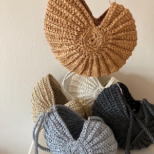 Könnte beinhalten: Eine Sammlung von gewebten Raffia-Handtaschen in verschiedenen Farben, darunter Beige, Grau und Schwarz. Die Taschen haben eine einzigartige muschelartige Form mit einem zentralen kreisförmigen Design. Jede Tasche hat einen geflochtenen Griff und einen Metallverschluss.