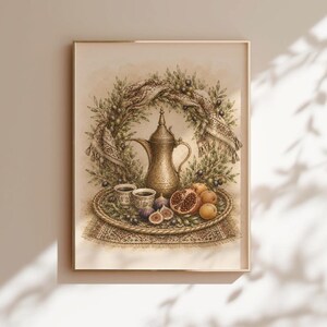May include: A framed art print featuring a detailed illustration of a metallic coffee pot, two small cups, a pomegranate, and oranges arranged on a woven tray. The scene is framed by a lush olive branch wreath with a patterned textile draped over it.