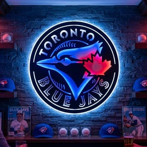 Pode incluir: Um letreiro de parede circular dos Toronto Blue Jays com um contorno LED brilhante em azul e branco. O logótipo apresenta uma cabeça de gaio-azul estilizada e uma folha de ácer vermelha. O letreiro está montado numa parede de pedra escura.