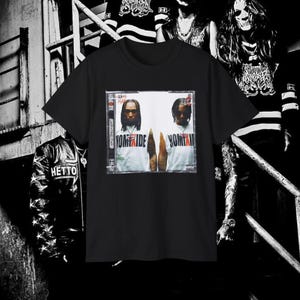 May include: Black t-shirt featuring a graphic of two people with the text "NOMIHIDE" and "HOMITII" on a white background. The shirt has a crew neck and short sleeves. The image is a close-up of the shirt.