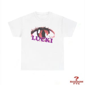May include: White t-shirt featuring a stylized eye graphic in shades of red and black, with the word "LUCKI" in purple. The design is centered on the front of the shirt. The shirt has a classic crew neck and short sleeves.