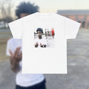 Op de afbeelding: Wit T-shirt met een afbeelding van een persoon, de tekst "Sybau" en een rood hart. De persoon maakt een handgebaar. Het T-shirt heeft een klassieke ronde hals.