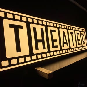 May include: Illuminated "THEATER" sign in a film strip design. The sign has a black border with white lettering and a warm, glowing light. The sign is mounted on a dark surface, creating a dramatic effect.