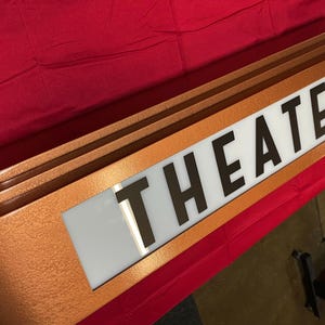 Puede incluir: Un letrero de marquesina de teatro de color cobre con la palabra "THEATER" en letras negras en negrita sobre un fondo blanco. El letrero tiene un dise&ntilde;o curvo y est&aacute; sobre un fondo rojo.
