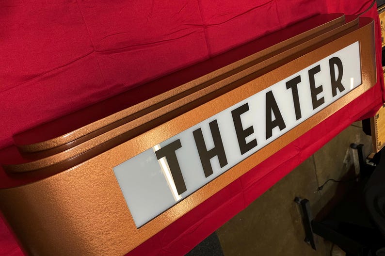 Puede incluir: Un letrero de marquesina de teatro de color cobre con la palabra "THEATER" en letras negras en negrita sobre un fondo blanco. El letrero tiene un dise&ntilde;o curvo y est&aacute; sobre un fondo rojo.