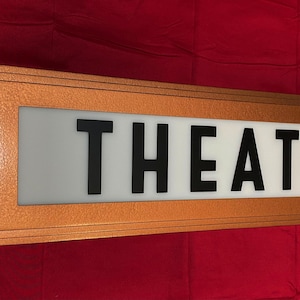 Puede incluir: Un letrero de marquesina "THEATER" vintage con un marco de metal color cobre. El letrero tiene un panel blanco con letras negras. El letrero rectangular tiene un dise&ntilde;o retro.