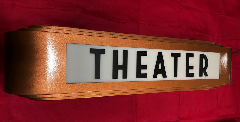 Puede incluir: Un letrero de marquesina "THEATER" vintage con un marco de metal color cobre. El letrero tiene un panel blanco con letras negras. El letrero rectangular tiene un dise&ntilde;o retro.