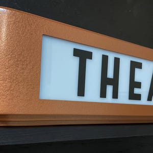 Puede incluir: Un letrero de marquesina de color cobre con la palabra "THEATER" en letras negras en negrita. El letrero tiene forma rectangular con bordes redondeados y un panel blanco iluminado. El letrero est&aacute; montado sobre una superficie oscura.