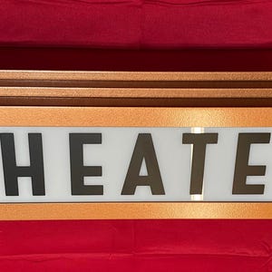 Puede incluir: Un letrero de teatro de estilo vintage con la palabra "THEATER" en letras negras. El letrero tiene un marco de color cobre y un fondo blanco para las letras. El letrero es rectangular con un dise&ntilde;o curvo.