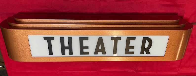 Puede incluir: Un letrero de teatro de estilo vintage con la palabra "THEATER" en letras negras. El letrero tiene un marco de color cobre y un fondo blanco para las letras. El letrero es rectangular con un dise&ntilde;o curvo.
