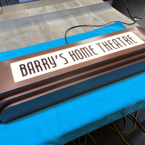 Puede incluir: Un letrero marr&oacute;n de estilo retro con el texto "BARRY'S HOME THEATRE" en letras blancas. El letrero tiene un dise&ntilde;o en capas y est&aacute; iluminado. Est&aacute; sobre una superficie azul.