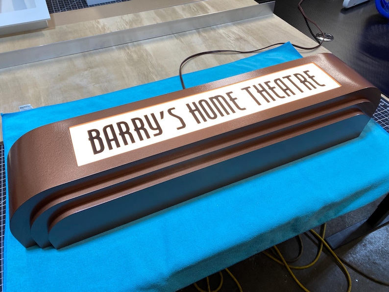 Puede incluir: Un letrero marr&oacute;n de estilo retro con el texto "BARRY'S HOME THEATRE" en letras blancas. El letrero tiene un dise&ntilde;o en capas y est&aacute; iluminado. Est&aacute; sobre una superficie azul.