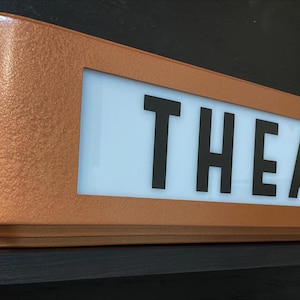 Puede incluir: Un letrero de marquesina "THEATER" de estilo retro, de color cobre. El letrero presenta un acabado texturizado y un panel blanco iluminado con letras negras en negrita. El dise&ntilde;o evoca una est&eacute;tica de cine vintage.