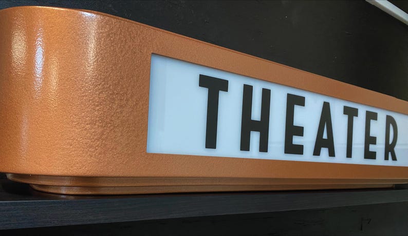 Puede incluir: Un letrero de marquesina "THEATER" de estilo retro, de color cobre. El letrero presenta un acabado texturizado y un panel blanco iluminado con letras negras en negrita. El dise&ntilde;o evoca una est&eacute;tica de cine vintage.