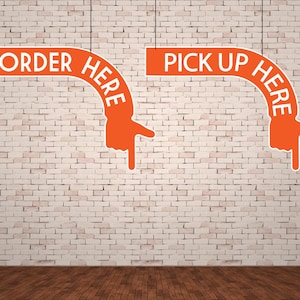 May include: Two orange signs with white text hang against a brick wall. One sign reads "ORDER HERE" and the other reads "PICK UP HERE". Both signs have a hand pointing downwards.
