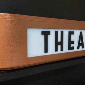 Puede incluir: Un letrero de teatro de color cobre con la palabra "THEATRE" en letras may&uacute;sculas negras y en negrita. El letrero tiene forma rectangular con bordes redondeados y est&aacute; montado sobre una superficie negra.