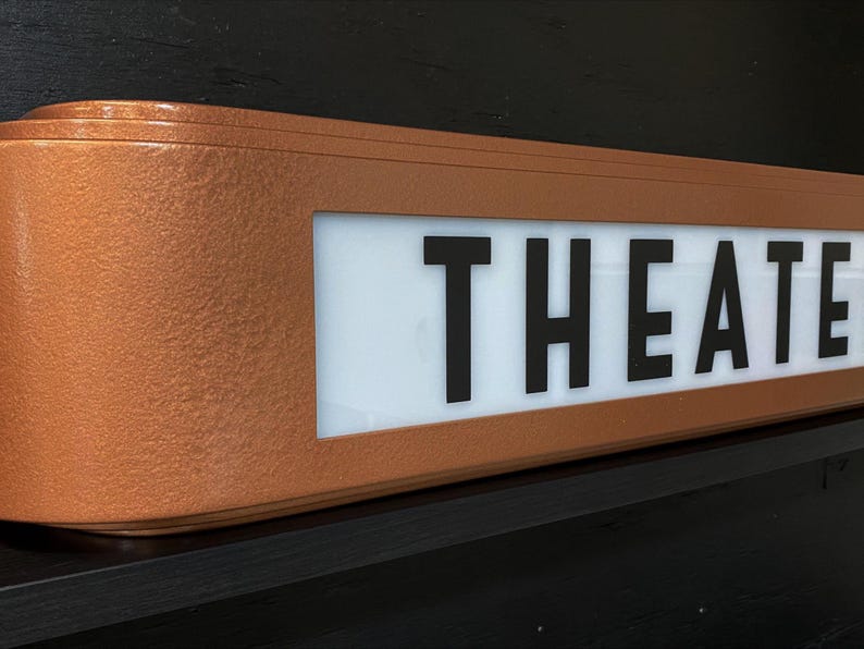 Puede incluir: Un letrero de teatro de color cobre con la palabra "THEATRE" en letras may&uacute;sculas negras y en negrita. El letrero tiene forma rectangular con bordes redondeados y est&aacute; montado sobre una superficie negra.
