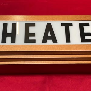 Puede incluir: Un letrero de teatro rectangular de color cobre con la palabra "THEATER" en letras negras en negrita sobre un fondo blanco. El letrero tiene un dise&ntilde;o en capas y un cable de alimentaci&oacute;n. Sobre una superficie roja.