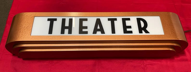 Puede incluir: Un letrero de teatro rectangular de color cobre con la palabra "THEATER" en letras negras en negrita sobre un fondo blanco. El letrero tiene un dise&ntilde;o en capas y un cable de alimentaci&oacute;n. Sobre una superficie roja.