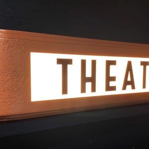 Puede incluir: Un letrero de teatro rectangular de color cobre con la palabra "THEATER" iluminada en blanco. El letrero tiene un dise&ntilde;o retro art d&eacute;co con bordes redondeados y un acabado texturizado. Est&aacute; montado sobre una superficie negra.
