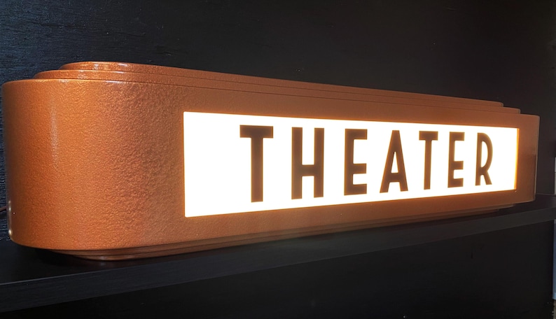 Puede incluir: Un letrero de teatro rectangular de color cobre con la palabra "THEATER" iluminada en blanco. El letrero tiene un dise&ntilde;o retro art d&eacute;co con bordes redondeados y un acabado texturizado. Est&aacute; montado sobre una superficie negra.