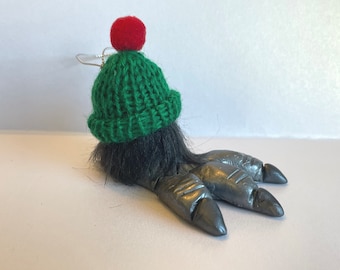May include: A green knitted hat with a red pom-pom on top, sitting on a gray, furry, three-toed foot ornament.