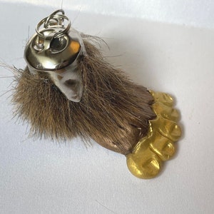 May include: A brown furry keychain with a gold foot and a silver ring. The keychain is shaped like a foot with four toes.