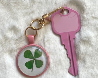 May include: A pink key chain with a gold metal ring and a round charm featuring a pressed four-leaf clover. The charm has a clear glass cover and a pink border. The key chain also includes a pink house key.
