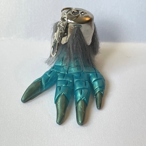 May include: A blue and silver monster claw keychain with a furry top. The claw has four sharp, pointed fingers.
