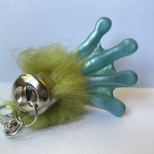 May include: A green and blue fuzzy frog keychain with a silver ring. The frog's foot is blue and the rest of the body is green and fuzzy.