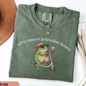 May include: A moss green t-shirt featuring a graphic of a frog wearing a bandana and sunglasses, riding a scooter while holding a shopping bag and a drink. The text "off to commit godmother foolery" is printed above the frog in white lettering.