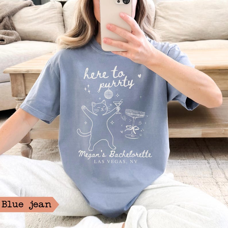 May include: Blue jean colored t-shirt with the text "here to purrty" and a graphic of a cat holding a martini. Below the graphic is "Megan's Bachelorette, Las Vegas, NV".