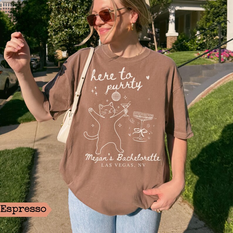 May include: Espresso-colored t-shirt with the text "here to purrty" and a graphic of a cat holding a cocktail. The shirt also says "Megan's Bachelorette, Las Vegas, NV".