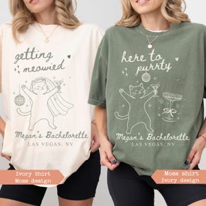 May include: Two t-shirts, one ivory and one moss green, with cat-themed bachelorette party designs. The ivory shirt says "getting meowied" and the green shirt says "here to purrty". Both shirts feature a cat illustration and "Megan's Bachelorette, Las Vegas, NV".