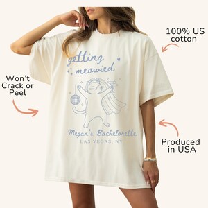 May include: Cream-colored oversized t-shirt with the phrase "getting meowied" and a cartoon cat in a veil, holding a disco ball and a cocktail. The text "Megan's Bachelorette Las Vegas, NV" is below the cat. Made of 100% US cotton.