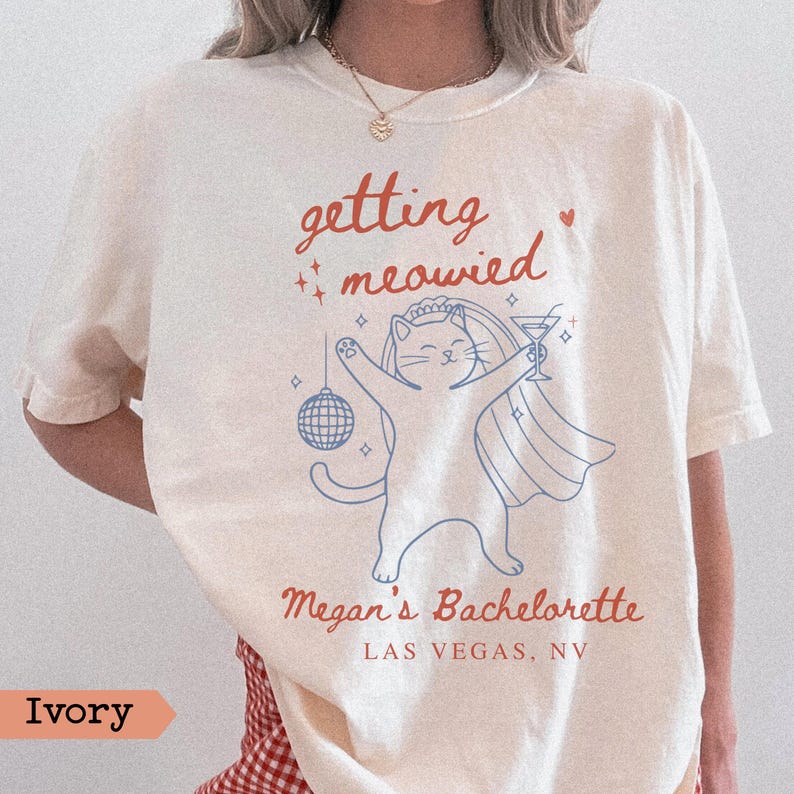 May include: Ivory-colored t-shirt with the phrase "getting meowied" in red script, a cartoon cat in a veil holding a martini and disco ball. The text "Megan's Bachelorette LAS VEGAS, NV" is below the cat illustration.