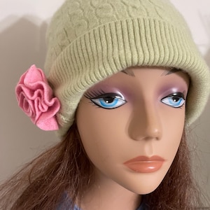 May include: A light green knit beanie with a cable knit pattern and a pink felt flower on the side. The hat is displayed on a mannequin head. The beanie is a stylish accessory.