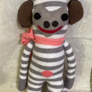 May include: A stuffed monkey toy crafted from grey and white striped knit fabric. It features dark brown ears and a tuft of hair on top, black circular eyes, a red stitched mouth, a pink scarf around its neck, and a small pink circle on its belly.